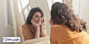 woman smiling at her reflection in a mirror to illustrate article about positive thinking