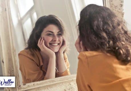 woman smiling at her reflection in a mirror to illustrate article about positive thinking